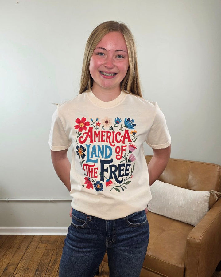 A smiling teenage girl with long blonde hair and braces stands indoors in front of a tan chair, wearing the Cosmos + Clover Boutique Land of the Free Graphic Tee. The shirt features colorful flowers and “America Land of the Free” text.