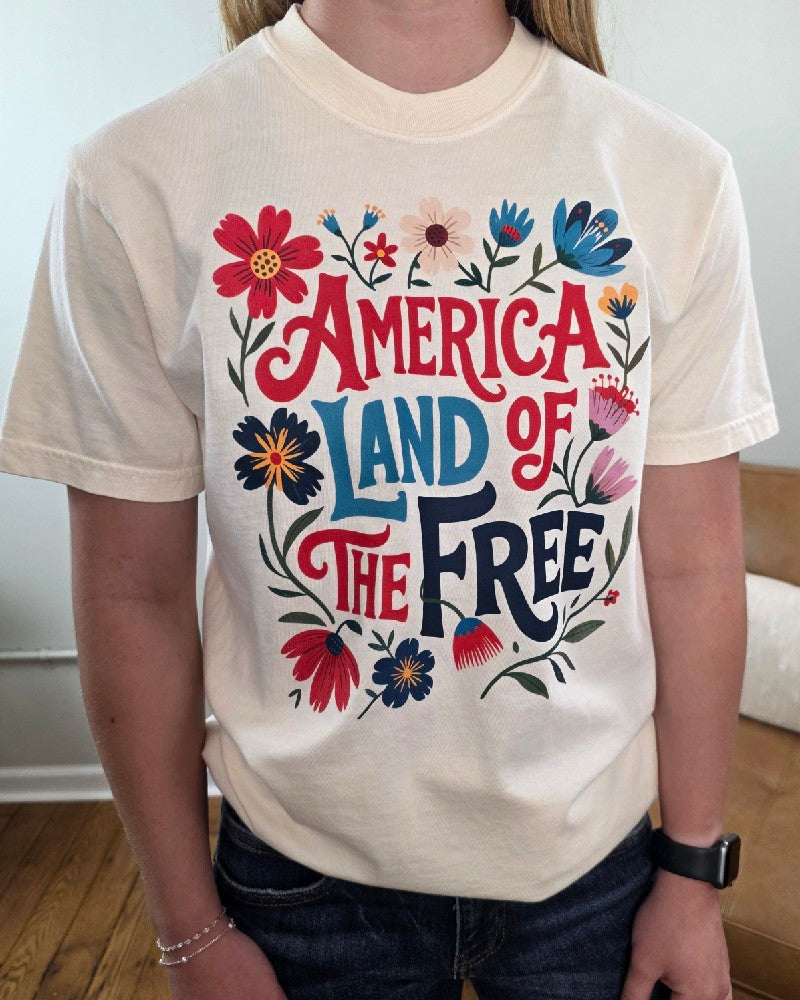 A person wearing the Land of the Free Graphic Tee by Cosmos + Clover Boutique, featuring colorful flowers and bold "America Land of the Free" lettering—a standout patriotic shirt.