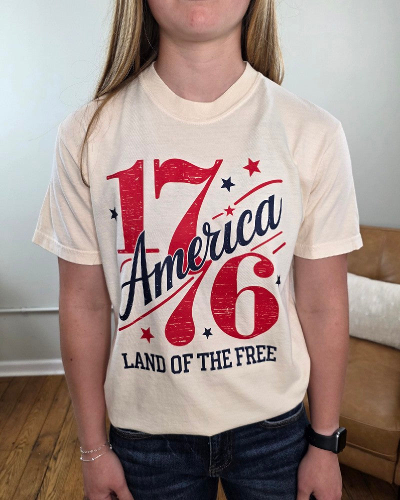 A person stands indoors wearing a cream-colored Cosmos + Clover Boutique Land of the Free 1776 Graphic Tee with "1776 America Land of the Free" in bold red and blue letters with stars. Only their torso and part of their arms are visible.