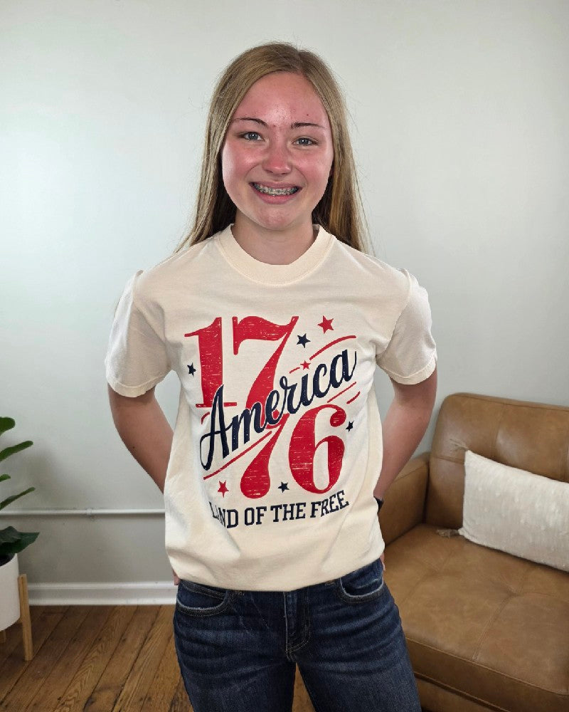 A smiling teenage girl with long blonde hair and braces wears the Land of the Free 1776 Graphic Tee from Cosmos + Clover Boutique, featuring red, white, and blue lettering. She stands indoors by a tan couch and plant, radiating American spirit.