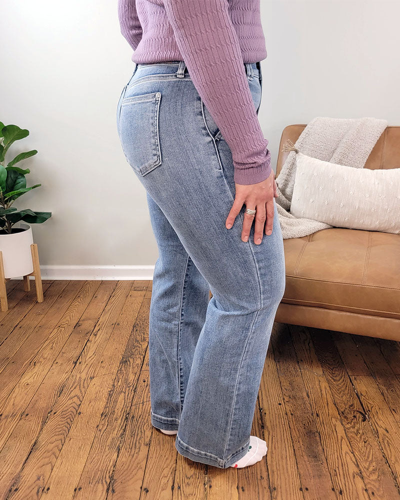 Someone wearing the Judy Blue Julia Wide Leg Jeans and a mauve long-sleeve top stands on a wooden floor near a tan couch with pillows and a plant in the background.