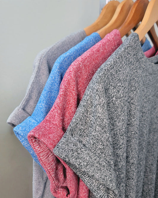 Four ZENANA Tania Tops in gray, blue, red, and dark gray hang on wooden hangers against a light background. The crew neck, short sleeve shirts have a soft heathered texture and are neatly arranged in a row.