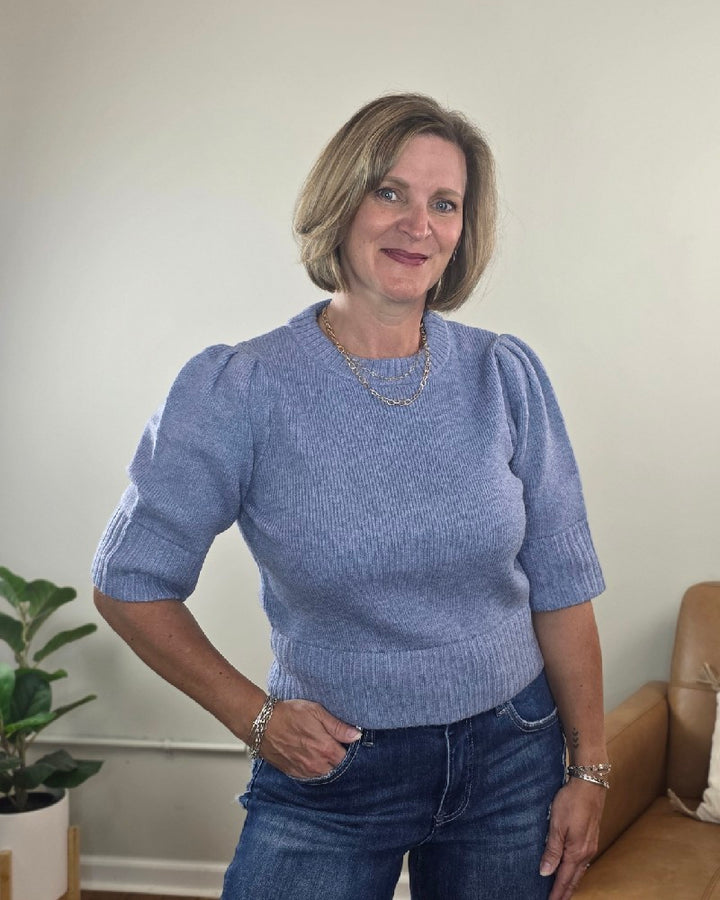 A woman with short blonde hair, wearing the blu pepper Lily Short Sleeve Pullover Sweater in light blue and jeans, stands indoors smiling with her hand in her pocket. A potted plant and a brown sofa are visible in the background.