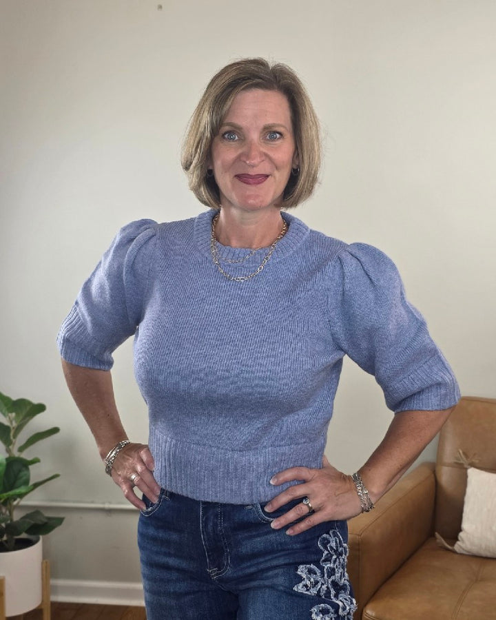 A woman with short blonde hair stands smiling indoors with her hands on her hips, wearing the blu pepper Lily Short Sleeve Pullover Sweater in light purple paired with floral embroidered jeans. A plant and a tan couch are visible in the background.