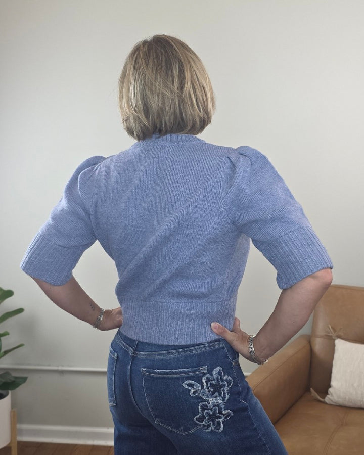A person with short blonde hair faces away, wearing the blu pepper Lily Short Sleeve Pullover Sweater in light blue and blue jeans with white floral embroidery on the back pocket.