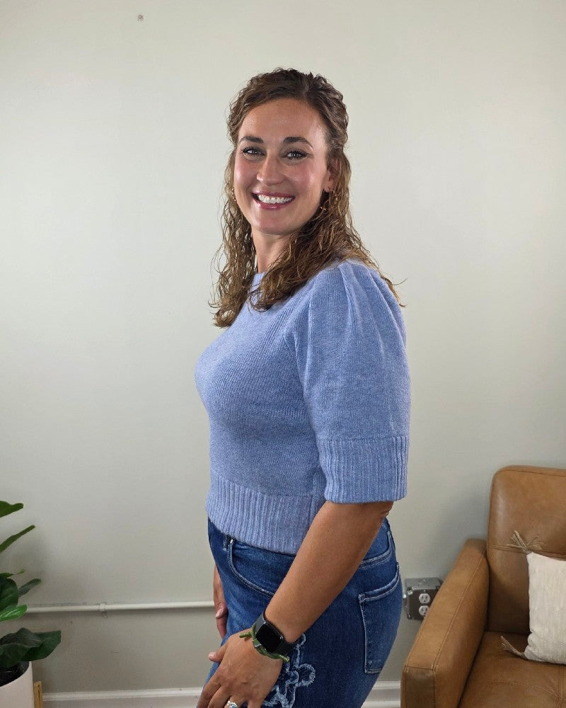 A woman with wavy brown hair smiles at the camera in a light-colored room with a plant and a brown chair, wearing the blu pepper Lily Short Sleeve Pullover Sweater in light blue paired with jeans.