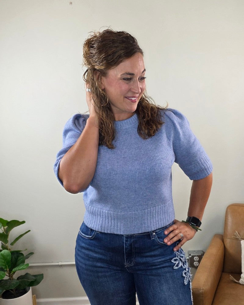 A smiling woman with curly hair stands indoors near a plant and a tan chair, wearing the blu pepper Lily Short Sleeve Pullover Sweater in light blue and jeans, with one hand touching her hair.