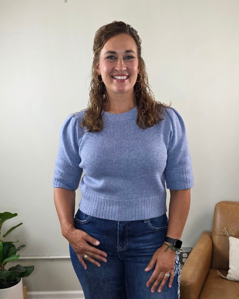 A woman with curly brown hair, wearing the blu pepper Lily Short Sleeve Pullover Sweater in light blue and dark jeans, stands smiling in a living room with a plant and a tan leather chair in the background.