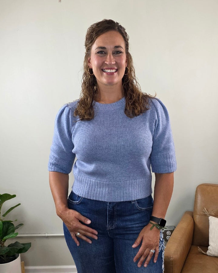 A woman with curly brown hair, wearing the blu pepper Lily Short Sleeve Pullover Sweater in light blue and dark jeans, stands smiling in a living room with a plant and a tan leather chair in the background.