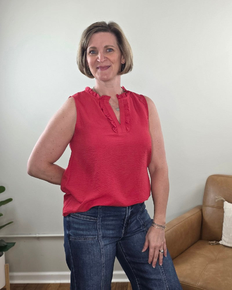 A woman with short light brown hair is smiling indoors, wearing the Staccato Lincoln Ruffled V-Neck Sleeveless Top and blue jeans. A tan armchair and a green potted plant are in the background.