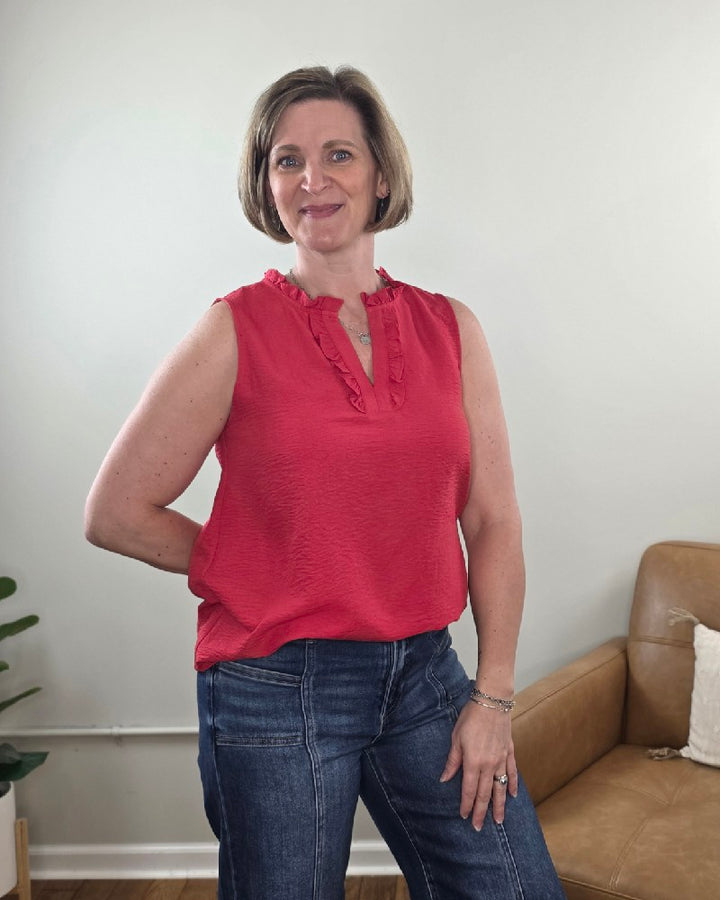 A woman with short light brown hair is smiling indoors, wearing the Staccato Lincoln Ruffled V-Neck Sleeveless Top and blue jeans. A tan armchair and a green potted plant are in the background.