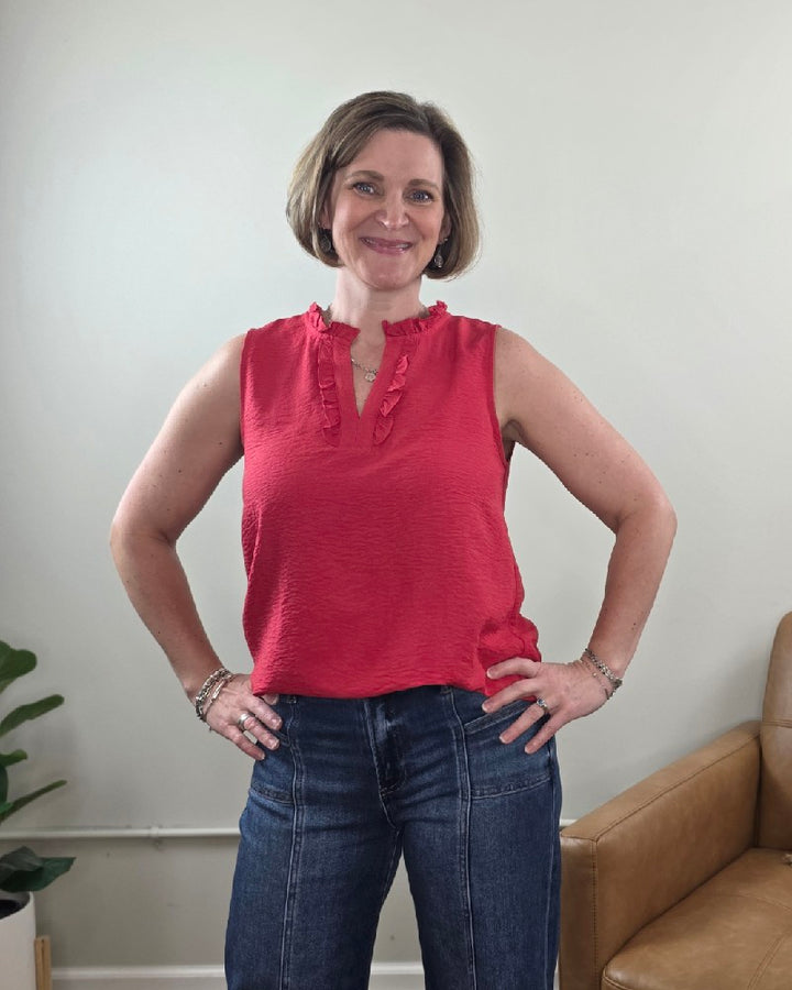 A woman with short hair smiles at the camera, standing with hands on hips in a red Staccato Lincoln Ruffled V-Neck Sleeveless Top and blue jeans. A green plant and tan chair are visible against a light-colored wall in the background.