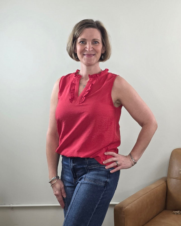 A woman with short brown hair smiles at the camera, wearing the Staccato Lincoln Ruffled V-Neck Sleeveless Top in red and blue jeans. She stands indoors next to a tan leather chair against a plain, light-colored wall.