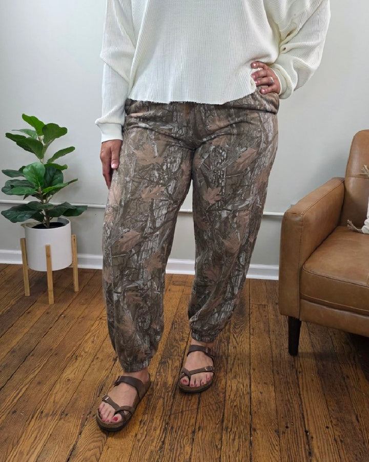 A person stands indoors wearing a white long-sleeve shirt, HYFVE, Inc Lottie Camo Joggers in a relaxed fit neutral print, and sandals. A green plant and brown armchair are in the background on a wooden floor.