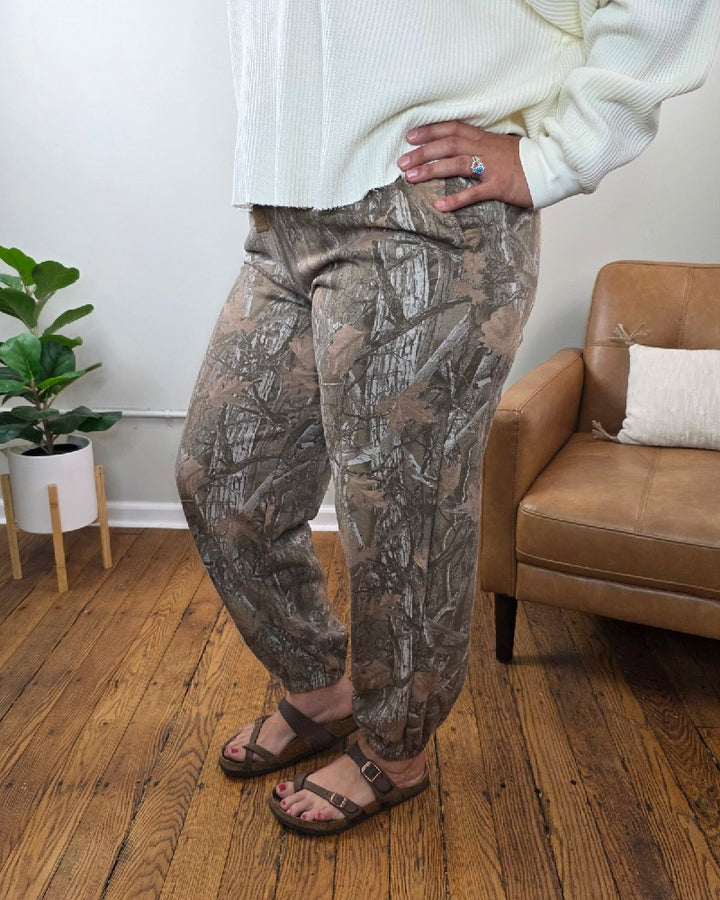 A person wearing HYFVE, Inc’s Lottie Camo Joggers pairs them with a cream sweater and brown sandals while standing on a wooden floor next to a tan chair and a potted plant. Only the lower half of their body is visible.