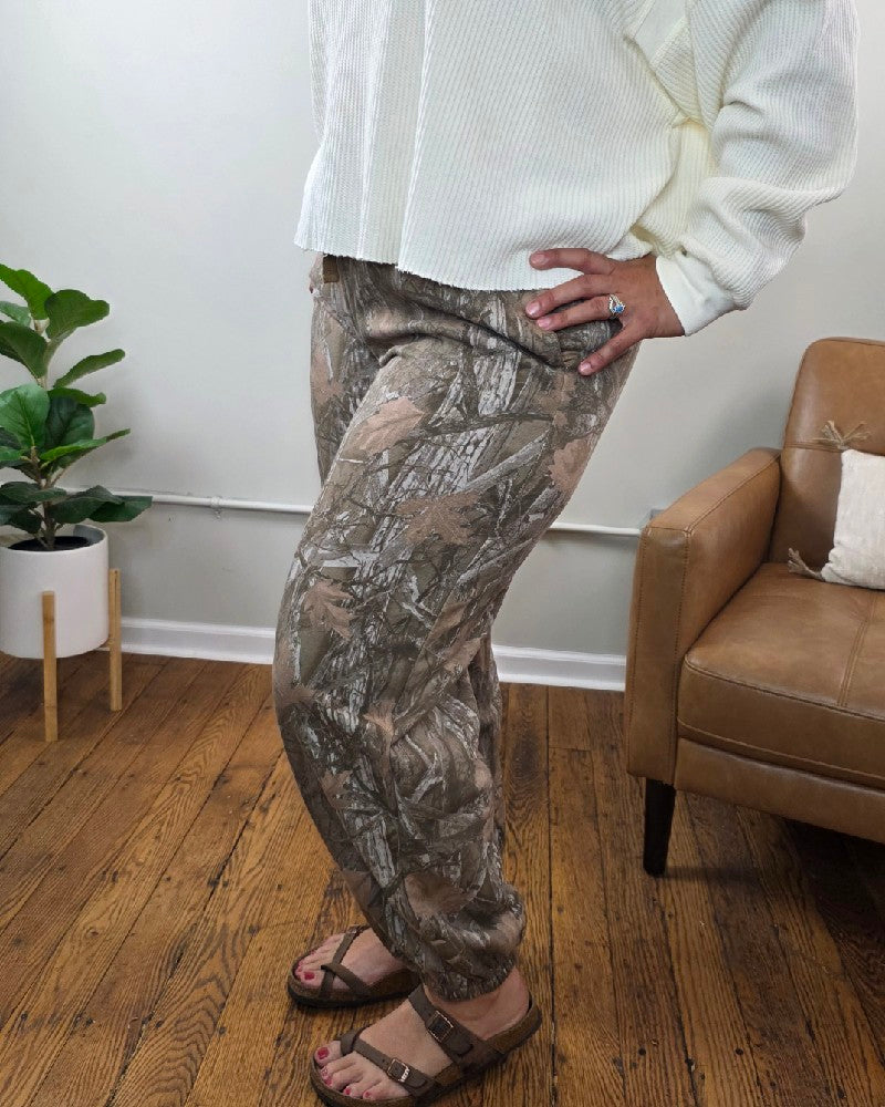 A person stands on a wood floor near a brown chair and potted plant, showcasing the Lottie Camo Joggers by HYFVE, Inc. paired with a cream textured sweater and brown sandals.