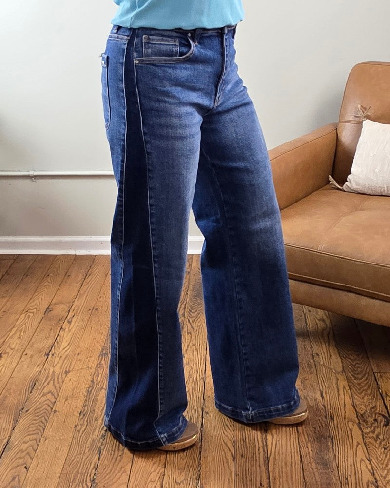 A person wearing the Mabel High Rise Wide Leg Contrast Jeans from Risen in a dark wash and brown shoes stands on a wooden floor next to a tan armchair with a white pillow, against a light-colored wall.