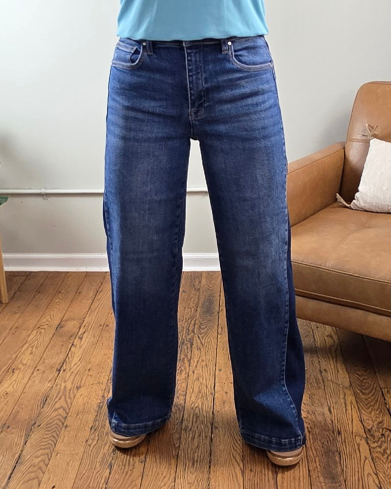 A person wearing the Mabel High Rise Wide Leg Contrast Jeans from Risen and brown shoes stands on a wooden floor next to a brown chair with a cushion.
