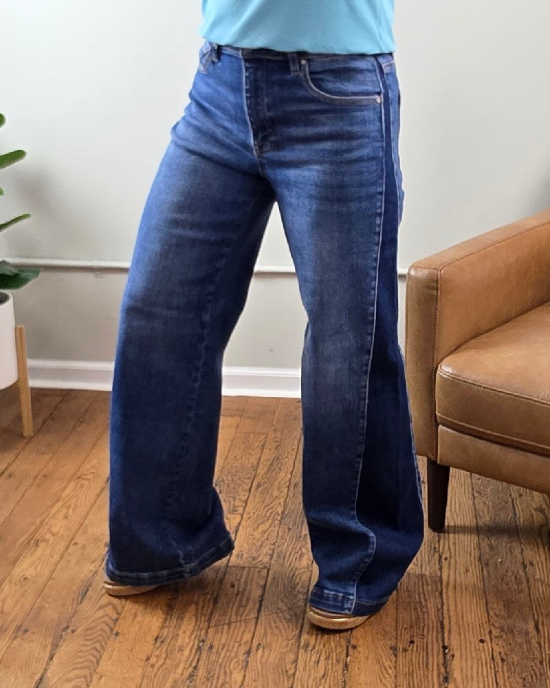 A person wearing the Risen Mabel High Rise Wide Leg Contrast Jeans, crafted from stretchy denim in blue, stands indoors on a wooden floor; the background features a brown armchair, a white wall, and a green potted plant.