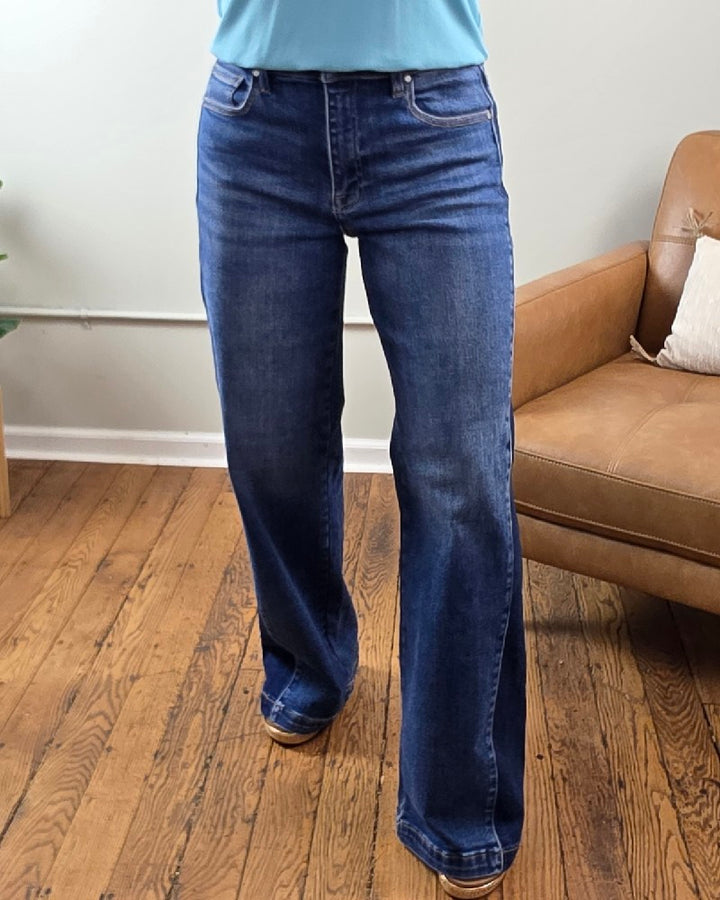 A person wearing the Risen Mabel High Rise Wide Leg Contrast Jeans, crafted from stretchy denim, and a light blue top stands on a wooden floor near a tan armchair and a plant, with a light-colored wall in the background.