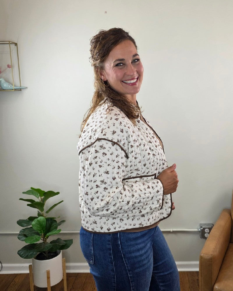 A woman with wavy brown hair smiles while standing sideways, wearing the blu pepper Mara Floral Button Down Jacket—perfect for fall layering—paired with blue jeans. Behind her are a green potted plant, a tan couch, and a white wall accented by a small shelf.