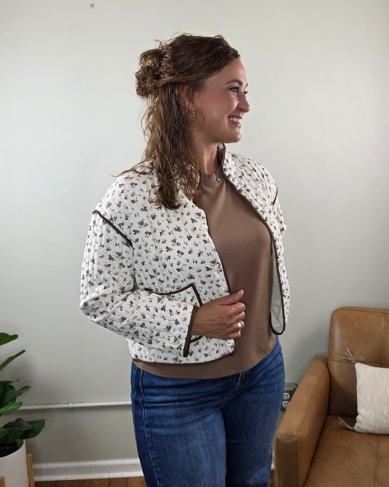 Wearing the Mara Floral Button Down Jacket by blu pepper over a brown shirt and blue jeans, she stands smiling with her wavy hair pulled back. Against a cozy backdrop featuring a tan chair and potted plant, she perfectly embodies effortless fall layering inspiration.