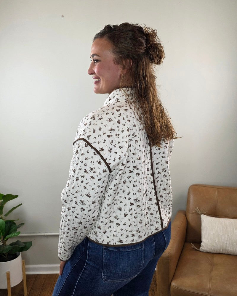 Woman with wavy brown hair wearing the blu pepper Mara Floral Button Down Jacket and blue jeans, standing and smiling in a modern living room with a tan couch and a potted plant—a perfect choice for fall layering.