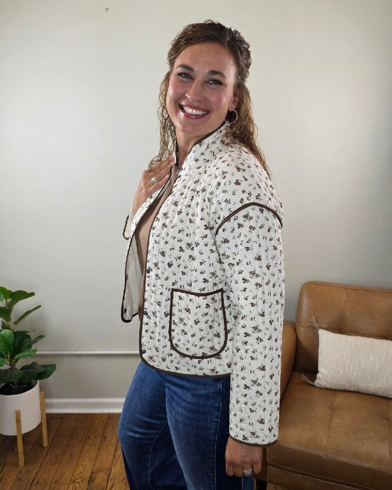 A smiling woman poses indoors wearing the Mara Floral Button Down Jacket by blu pepper, a light-colored quilted piece with brown trim, paired with blue jeans. A tan sofa and a green potted plant are in the background, accentuating this charming fall jacket that’s perfect as a lightweight layering piece.