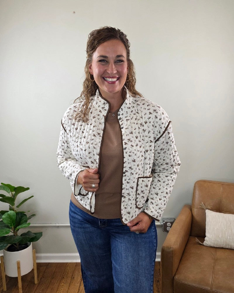 A woman with curly hair smiles as she wears the blu pepper Mara Floral Button Down Jacket paired with blue jeans. She stands indoors near a brown leather chair and a potted plant, set against a light-colored wall. The Mara Floral Button Down Jacket is a stylish and lightweight layering piece, perfect for cooler fall days.