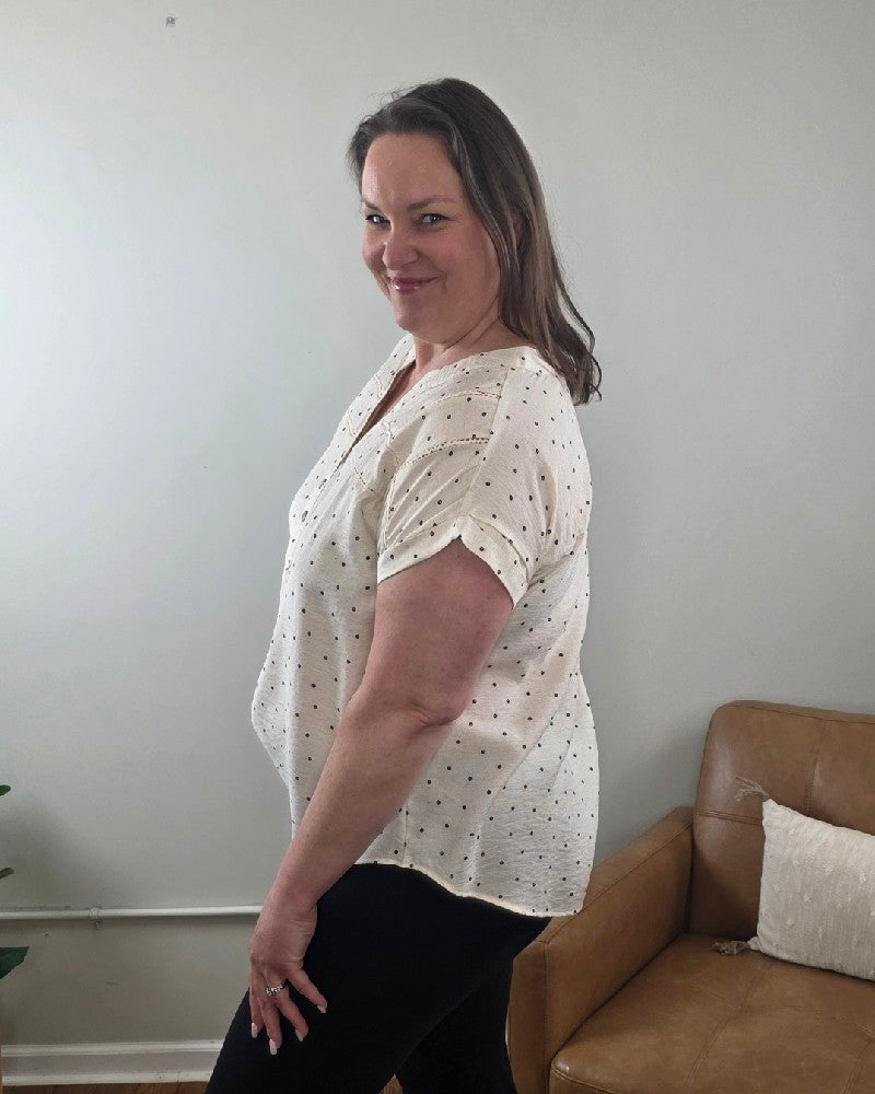 A woman with shoulder-length brown hair smiles as she stands sideways in a living room, wearing the Marin Polka Dot Lace Trim Button Top from Mine and E&M with black pants.