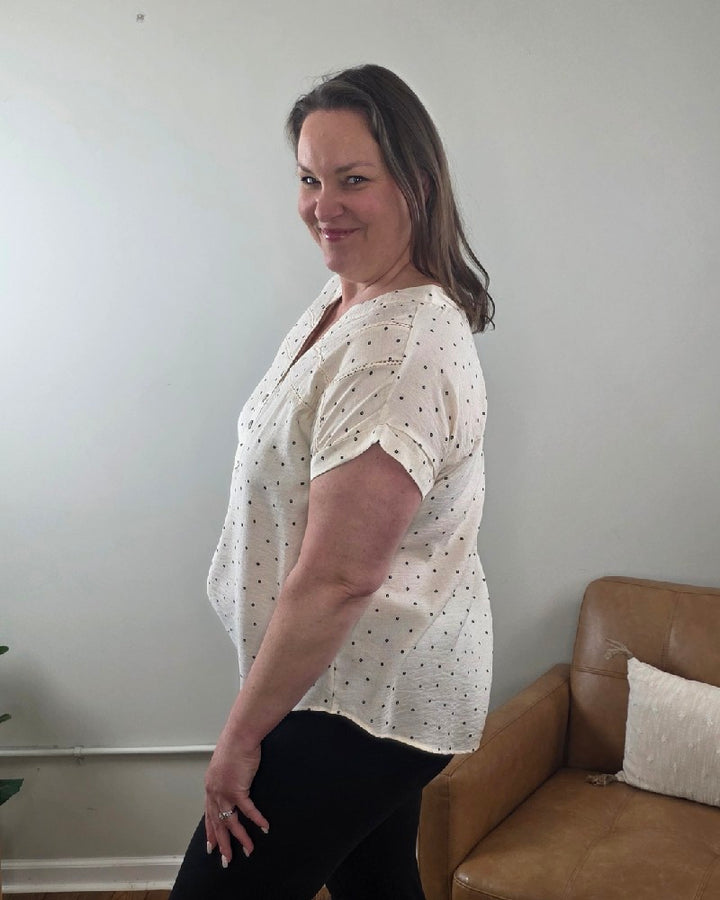 A woman with shoulder-length brown hair smiles as she stands sideways in a living room, wearing the Marin Polka Dot Lace Trim Button Top from Mine and E&amp;M with black pants.