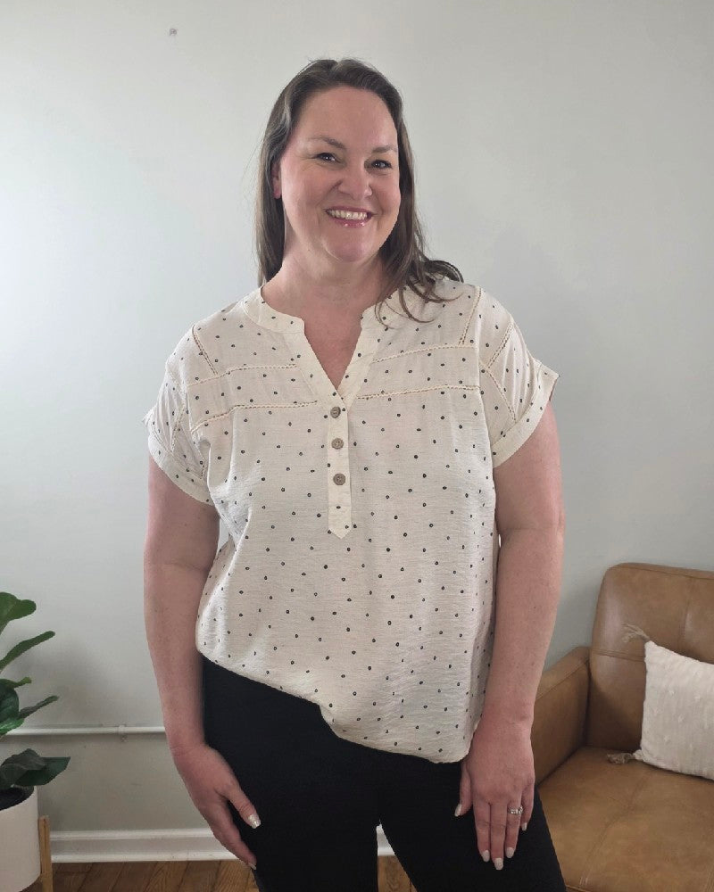 A woman with long brown hair smiles indoors, wearing the Marin Polka Dot Lace Trim Button Top by Mine and E&M and black pants. Behind her are a tan sofa, a cushion, and a green potted plant.