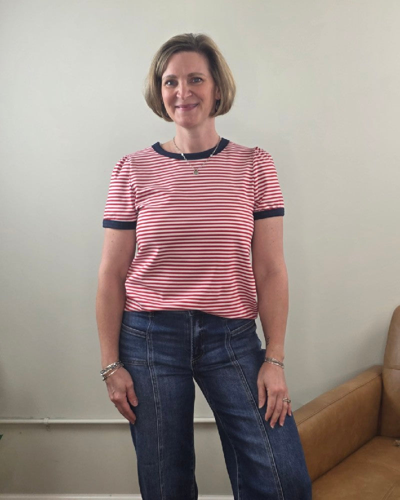 A woman with short brown hair stands indoors, smiling. She is wearing the Staccato Martha Color Trimmed, Short Sleeve, Striped Top and blue jeans. A tan couch sits nearby against a light-colored wall.