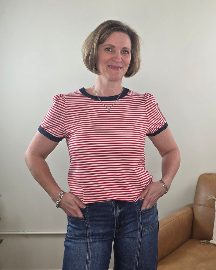 A woman with short brown hair smiles indoors, wearing the Staccato Martha Color Trimmed, Short Sleeve, Striped Top and blue jeans. Her hands are on her hips, and a tan couch is in the background.