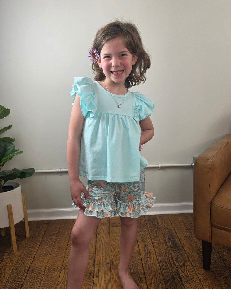 A young girl with shoulder-length brown hair and a flower in her hair smiles indoors, modeling the Adorable Sweetness Kids Miella Floral Summer Ruffle Short Set—an airy summer outfit for girls—standing on a wooden floor near a plant and brown armchair.