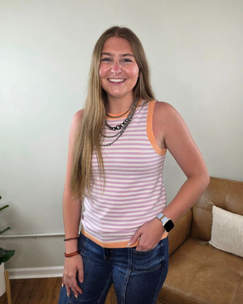 A young woman with long brown hair smiles indoors, wearing the Staccato Maybelle Color Contrast Striped Sleeveless Knit Top, layered necklaces, jeans, a smartwatch, and bracelets. Behind her are a tan couch and a plant against a light wall.