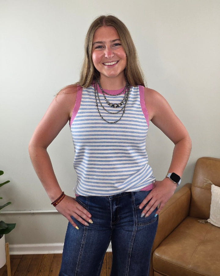 A smiling woman with long hair wears the Staccato Maybelle Color Contrast Striped Sleeveless Knit Top, layered necklaces, blue jeans, and a smartwatch as she stands indoors near a tan couch and a plant.
