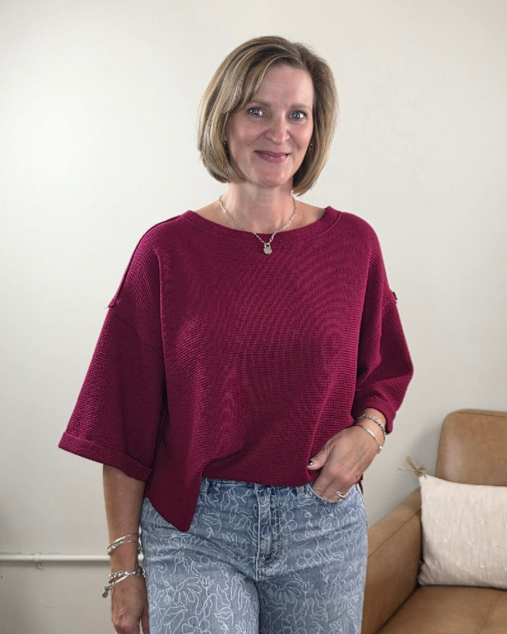 A woman with short blonde hair is seen smiling in a room with light walls and a brown couch, effortlessly capturing casual style in the ZENANA Melissa 3/4 Sleeve Waffle Knit Top paired with patterned blue jeans.