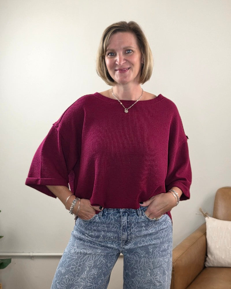 A woman with short blonde hair is standing and smiling in a light-colored room with a brown chair in the background, showcasing a relaxed, casual look in the ZENANA Melissa 3/4 Sleeve Waffle Knit Top paired with patterned blue jeans.