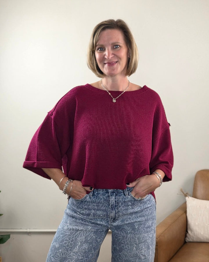 A woman with short blonde hair is standing and smiling in a light-colored room with a brown chair in the background, showcasing a relaxed, casual look in the ZENANA Melissa 3/4 Sleeve Waffle Knit Top paired with patterned blue jeans.