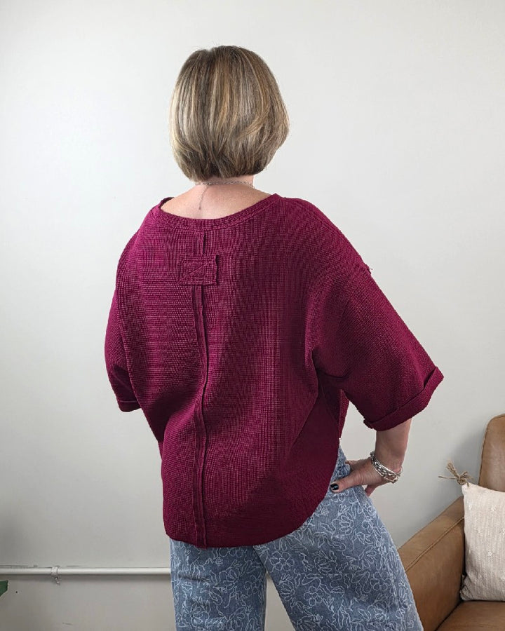 A person with short blond hair is standing with their back to the camera, wearing the ZENANA Melissa 3/4 Sleeve Waffle Knit Top in maroon and light blue patterned pants. This comfortable, relaxed outfit is part of a casual wardrobe and is seen in a room with white walls and a brown couch.