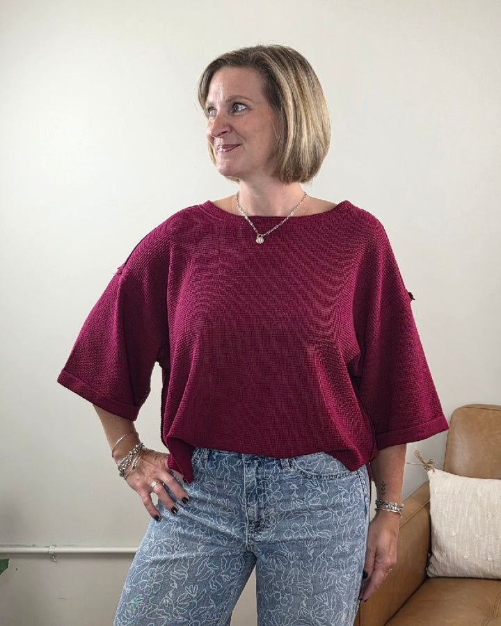 A woman with short hair wears the Melissa 3/4 Sleeve Waffle Knit Top by ZENANA in maroon, paired with patterned blue jeans. She stands indoors near a tan sofa, smiling and looking to her left—a perfect addition to any casual wardrobe.