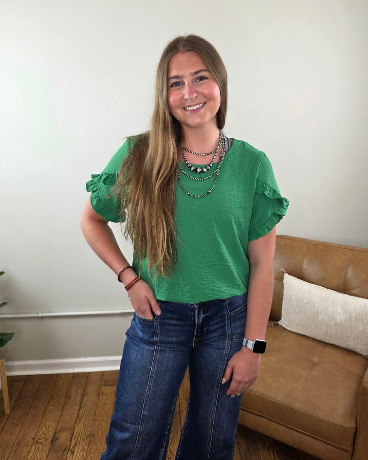 A woman with long brown hair smiles in a living room, wearing the Shewin Mellisa Ruffled Tulip Sleeve Blouse in kelly green and blue jeans. She accessorizes with layered necklaces, a smartwatch, and stands by a tan couch on a wooden floor.