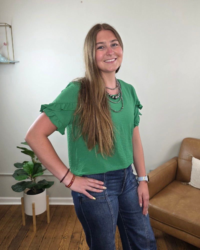A woman with long brown hair smiles in a living room with a brown sofa and potted plant, wearing the Shewin Mellisa Ruffled Tulip Sleeve Blouse in kelly green and jeans.