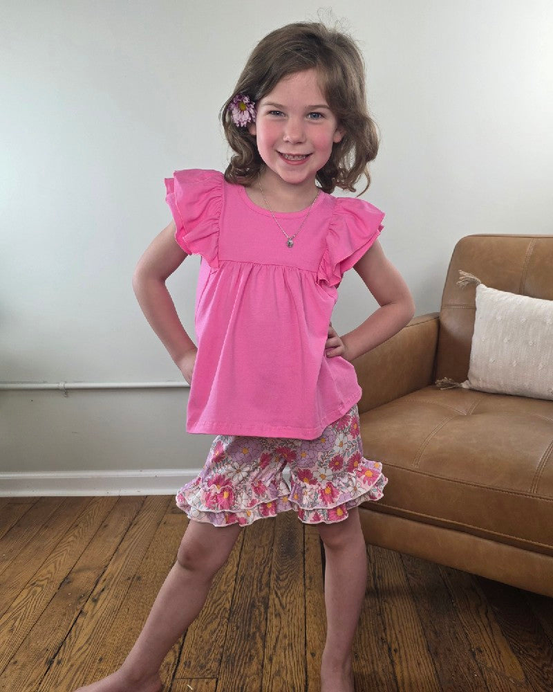 A young girl with brown hair smiles confidently indoors, wearing the Adorable Sweetness Kids Miella Floral Summer Ruffle Short Set—a stylish pink top with ruffled sleeves and floral shorts—perfect for girls' summer outfits beside a tan couch on wooden floors.