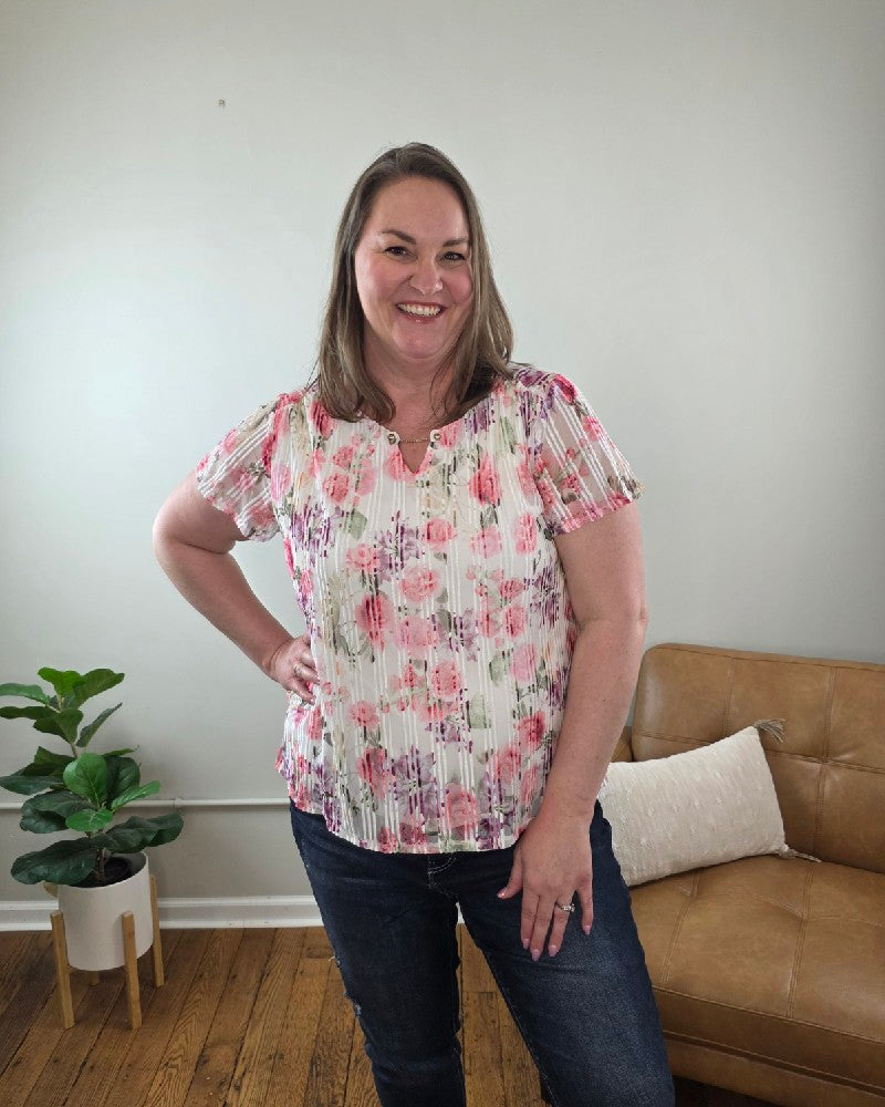 A woman with straight brown hair smiles indoors, wearing the Miranda Floral Print, Flutter Sleeve Blouse and jeans from Cosmos + Clover Boutique. She stands with one hand on her hip in front of a tan couch, pillow, green potted plant, and light-colored wall.