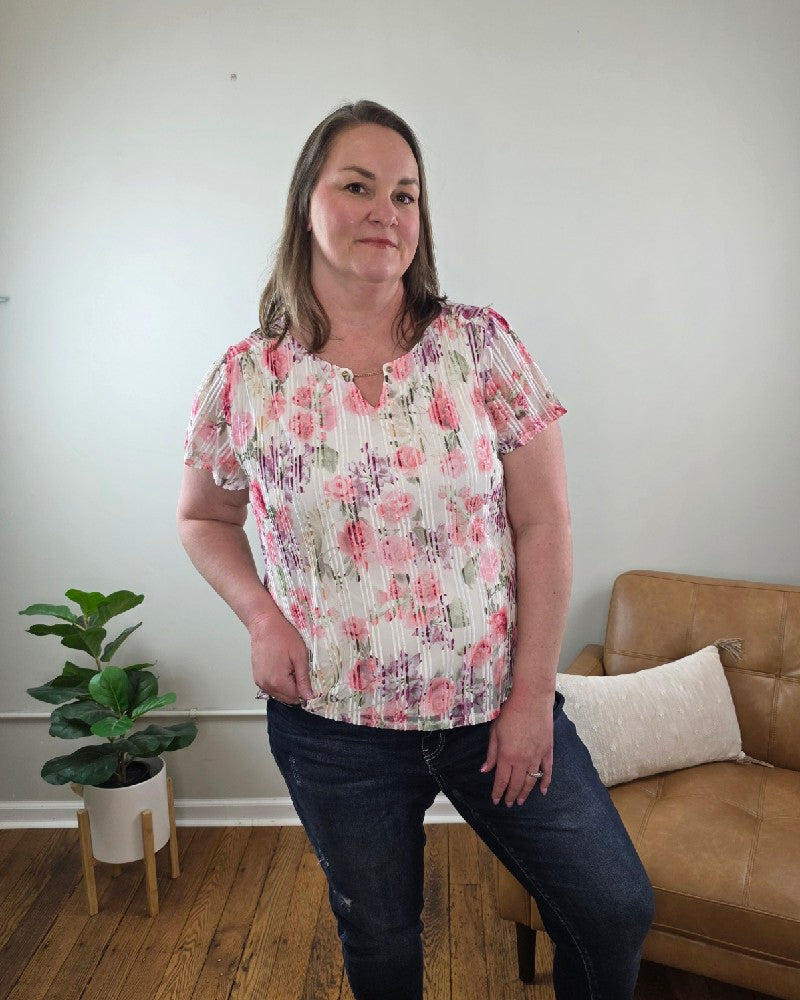 A woman with straight brown hair smiles indoors, wearing the Miranda Floral Print, Flutter Sleeve Blouse and jeans from Cosmos + Clover Boutique. She stands with one hand on her hip in front of a tan couch, pillow, green potted plant, and light-colored wall.