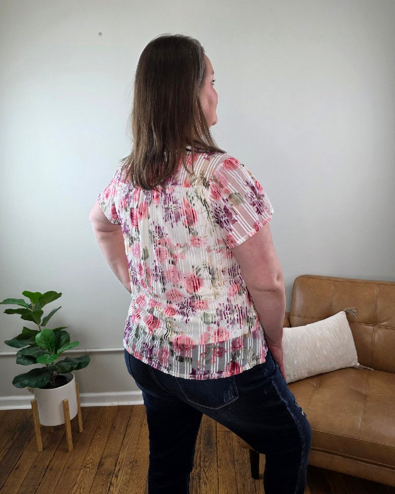 A woman stands indoors facing away from the camera, wearing the Miranda Floral Print Flutter Sleeve Blouse and dark jeans, beside a tan couch with a white pillow and a potted plant on the wooden floor.