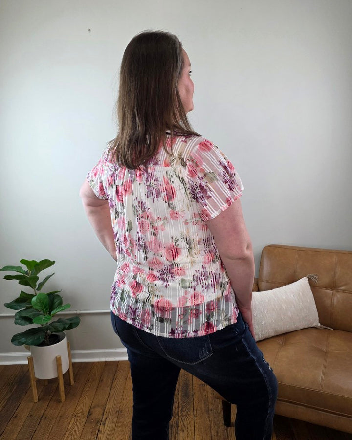 A woman stands indoors facing away from the camera, wearing the Miranda Floral Print Flutter Sleeve Blouse and dark jeans, beside a tan couch with a white pillow and a potted plant on the wooden floor.