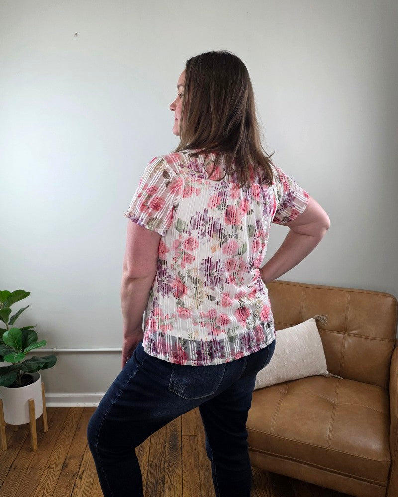 A woman with straight brown hair smiles indoors, wearing the Miranda Floral Print, Flutter Sleeve Blouse and jeans from Cosmos + Clover Boutique. She stands with one hand on her hip in front of a tan couch, pillow, green potted plant, and light-colored wall.